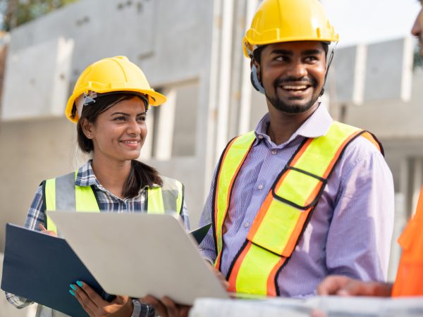 Team of construction engineers wearing vest and helmet safety discussing project at construction site.Group Indian foreman with laptop, paperwork working at factory marking precast concrete wall.